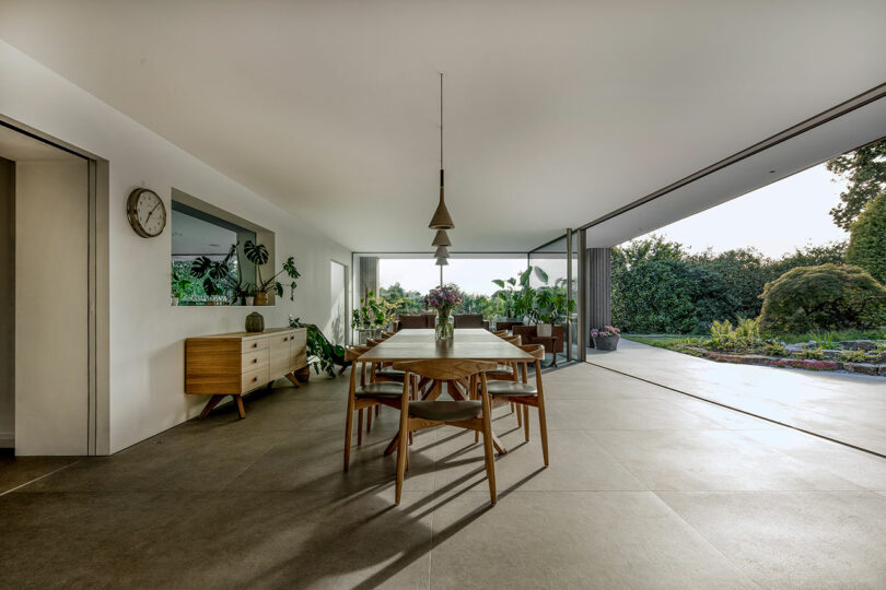 Spacious dining area with a wooden table, chairs, and pendant light. Large windows open to a garden view. Minimalist decor with plants on a sideboard.