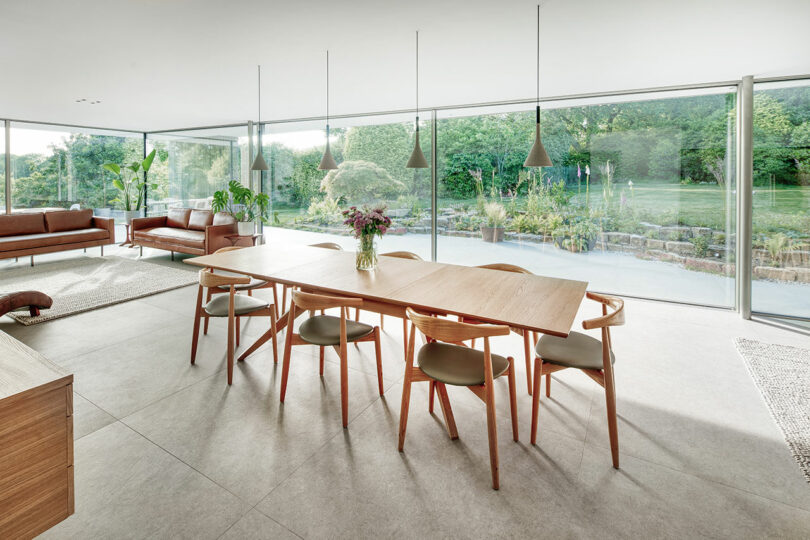 Minimalist dining area with a wooden table, chairs, and pendant lights. Large windows offer a view of a lush garden. Brown sofas and a rug are visible in the background.