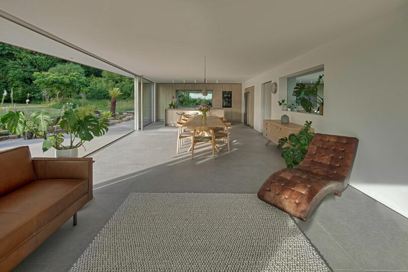 Modern open living space with a brown leather chair, sofa, and dining table. Floor-to-ceiling glass doors open to a green outdoor area. Neutral tones and indoor plants accent the room.