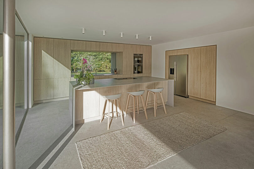 A modern kitchen with a central island, three wooden stools, and built-in appliances. Light wooden cabinets and a large window provide a spacious, airy feel. A rug is on the floor.