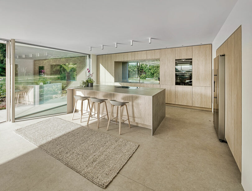 Modern kitchen with light wood cabinetry, large central island with stools, and floor-to-ceiling windows overlooking a garden. A textured rug lies on the floor.