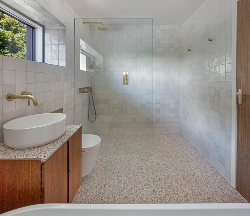 Modern bathroom with a glass-enclosed shower, white tile walls, a wooden vanity with an oval sink, and a window providing natural light. The shower features a gold rainfall showerhead.