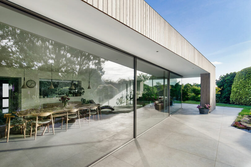 Modern glass-walled home with a dining area visible inside, surrounded by greenery and a clear sky outside.