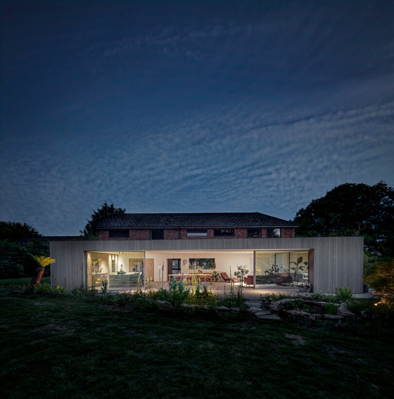 A modern two-story house with large windows is illuminated at night, surrounded by trees and a lawn under a partially cloudy sky.