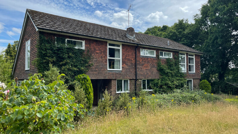 A two-story house with brown shingles partially covered in greenery, surrounded by overgrown grass and plants under a cloudy sky.