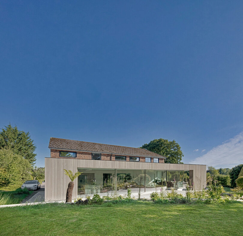 Modern house with large glass windows, wood accents, and a car parked beside it. Surrounded by grass and trees under a clear blue sky.