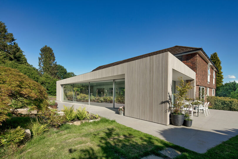 Modern extension on a traditional house with large glass windows, surrounded by greenery under a clear blue sky.