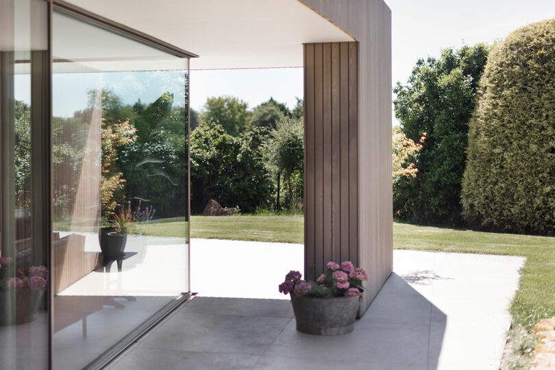 A modern building with large glass windows, a wooden exterior, and potted pink flowers on a concrete patio. Lush greenery surrounds the area.