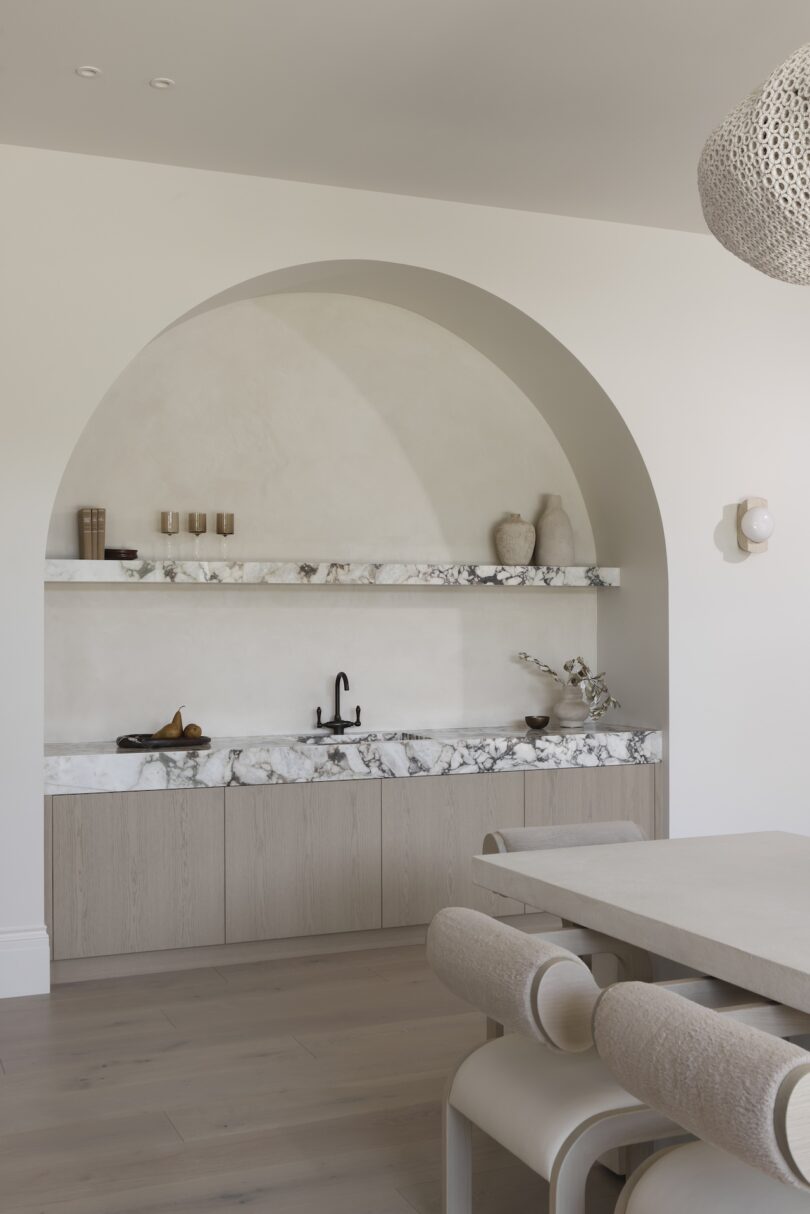 Minimalist kitchen with a marble countertop, arched alcove, open shelves with decor, a black faucet, and light wood cabinetry.