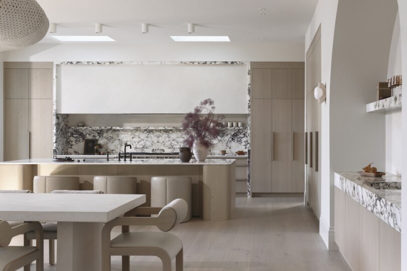 Modern kitchen with light wood cabinetry, marble countertops, and central island. Decorative vase with dried branches on the island. Minimalist dining table and chairs in foreground.