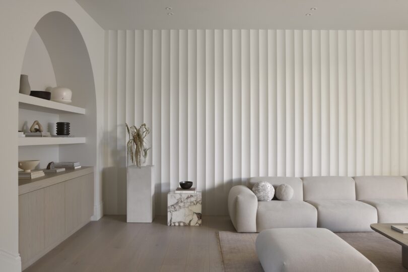 Modern living room with a textured white wall, beige sectional sofa, marble pedestal with decorative items, and built-in shelves holding vases and books. Neutral tones and minimalist design.