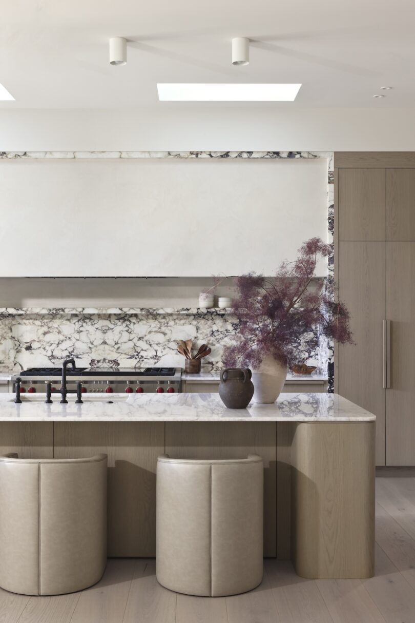 Modern kitchen with light wood cabinetry, marble backsplash, and a central island featuring two neutral-toned stools. A vase with purple foliage decorates the countertop.