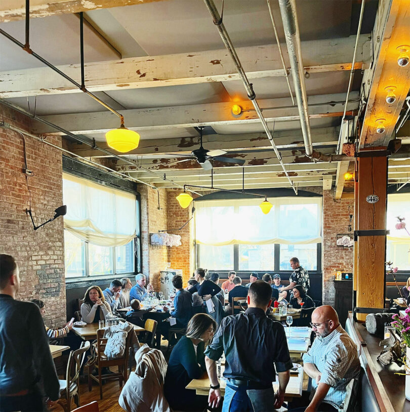 The bustling, rustic restaurant, designed by Chuck Cerankosky, features diners enjoying meals at wooden tables beneath exposed beams and industrial lighting. Large windows flood the space with natural light