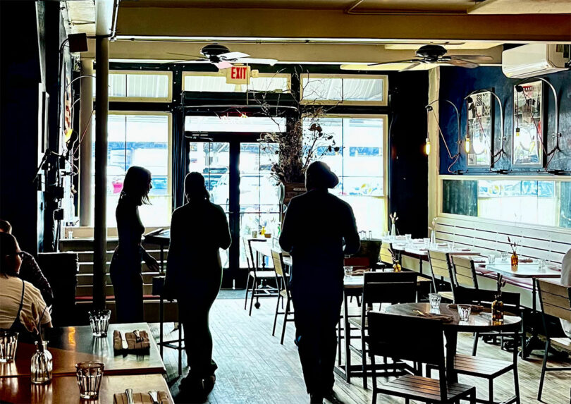 Three people meander through a dimly lit restaurant filled with tables and chairs, making their way toward the entrance