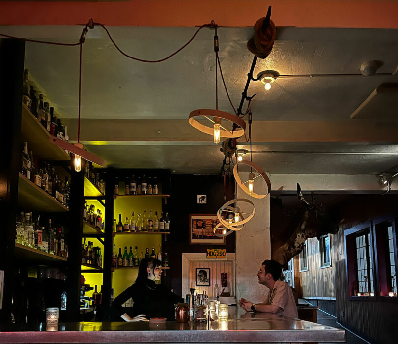 a dimly lit bar, shelves filled with bottles glimmer softly under the glow of circular overhead light fixtures.